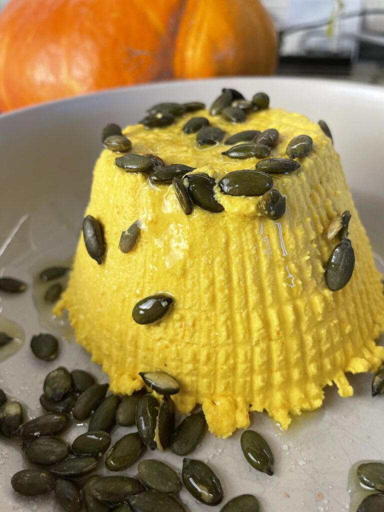 Veganer Ricotta aus Kürbis und Mandeln, gestürzt auf einem Teller mit gerösteten Kürbiskernen und Olivenöl